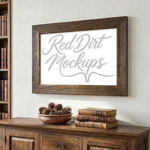 Könnte beinhalten: Ein rechteckiger gerahmter Mockup mit dem Text "Red Dirt Mockups" in einer Schreibschrift. Der Rahmen besteht aus dunkelbraunem Holz und hängt an einer weißen Wand. Darunter befindet sich ein Holzschrank mit einer Schale mit Tannenzapfen und gestapelten Büchern.