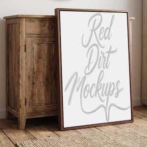 Peut inclure: Maquette de toile encadrée avec le texte "Red Dirt Mockups" en écriture cursive. Le cadre est marron foncé et la toile est blanche. La maquette est appuyée contre une armoire en bois sur un plancher en bois.
