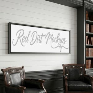 Puede incluir: Un lienzo enmarcado con el texto "Red Dirt Mockups" en cursiva. El marco es negro y el lienzo cuelga de una pared blanca con tablones. Dos sillones de cuero están frente a la pared y una estantería a la derecha.