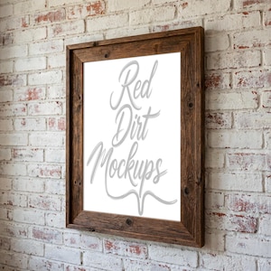 Könnte beinhalten: Ein rechteckiger Holzbilderrahmen mit dem Text "Red Dirt Mockups" in Schreibschrift. Der Rahmen besteht aus dunkelbraunem Holz und ist an einer weißen Backsteinmauer befestigt.