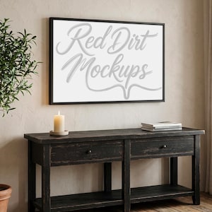 Op de afbeelding: Een ingelijst canvas met de tekst "Red Dirt Mockups" in een cursief lettertype hangt boven een donkere houten console. Een brandende kaars en een stapel boeken staan op de tafel. Een plant in een pot is links zichtbaar.