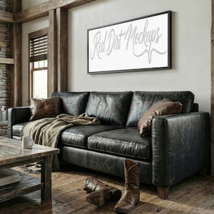 May include: A black leather sofa with two brown leather throw pillows and a tan blanket. A wooden coffee table and a pair of brown cowboy boots are in front of the sofa. A framed sign with the text "Red Dirt Mockups" hangs above the sofa.