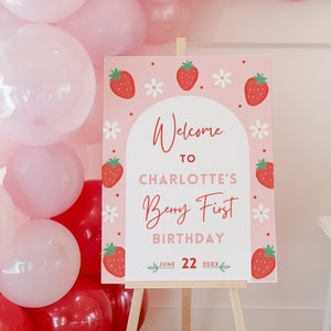 May include: A pink sign on a wooden easel with the text "Welcome to Charlotte's Berry First Birthday" and the date June 22, 20XX. The sign is decorated with strawberries and flowers. Pink, red, and green balloons are in the background.
