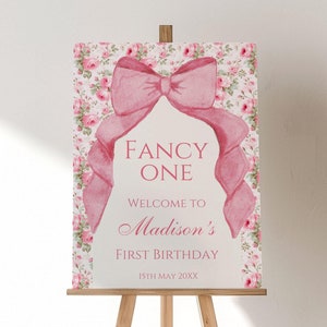 Op de afbeelding: Een roze en wit printbaar bord met een grote roze strik en de tekst "Fancy One"  "Welkom bij Madisons eerste verjaardag" en "15th May 20XX".