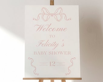 May include: A white sign with pink ribbon designs and the text "Welcome to Felicity's Baby Shower - 12" on an easel.