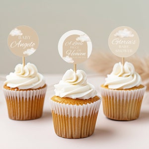 Könnte beinhalten: Drei Cupcakes mit weißem Frosting und braunen Cupcake-Förmchen. Jeder Cupcake hat einen runden, beigefarbenen Papier-Topper mit weißem Text. Die Topper lauten "Baby Angelo", "A Little Piece of Heaven" und "Gloria's Baby Shower".