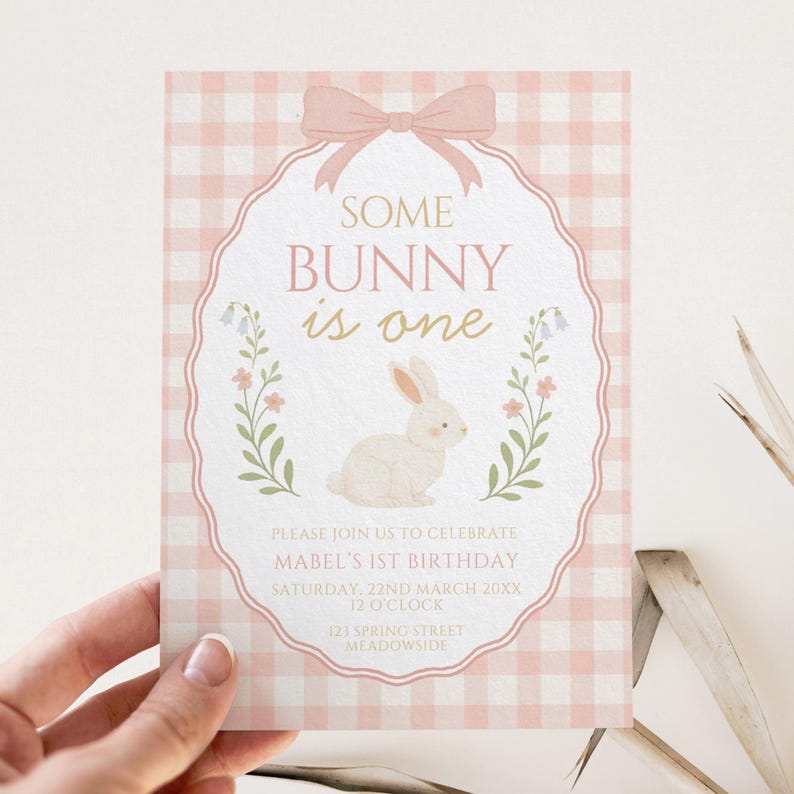 Op de afbeelding: Een roze en witte ruitjes verjaardag uitnodigingskaart met de tekst "SOME BUNNY is one". De kaart heeft een konijn illustratie en bloemmotieven, met details voor een eerste verjaardag.