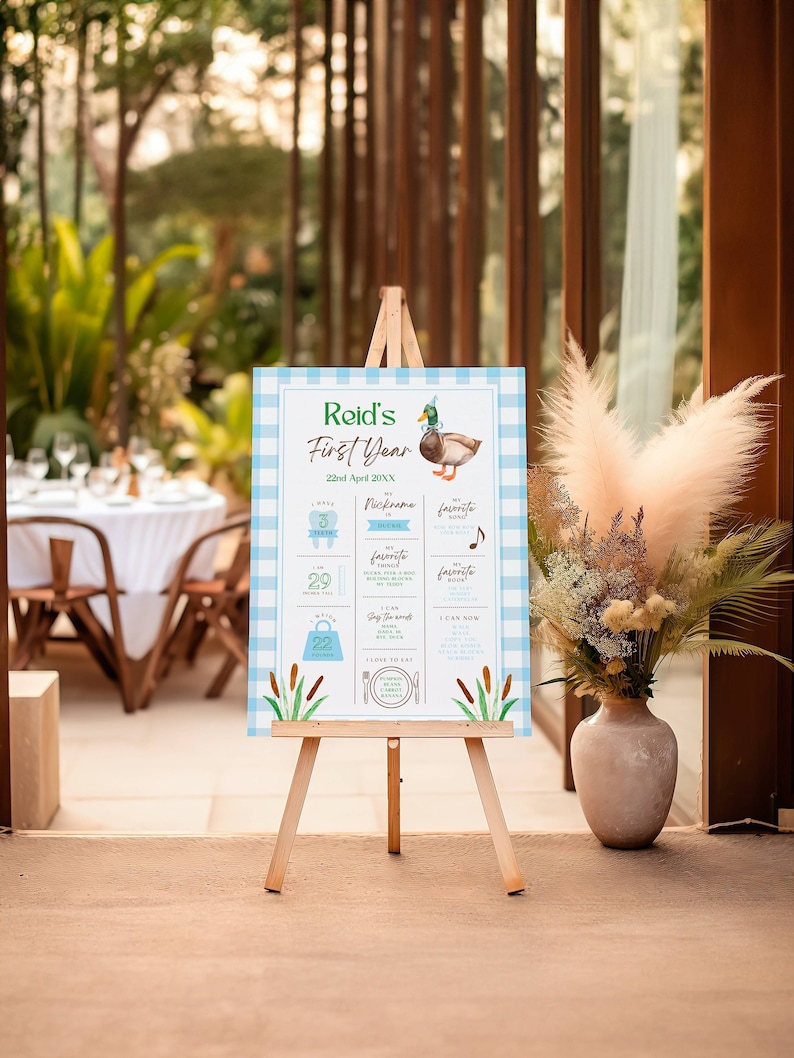 May include: A blue and white checkered poster with a duck illustration. The poster is on an easel and has text that says "Reid's First Year" and includes details about the child's first year, such as their weight, height, and favorite things.