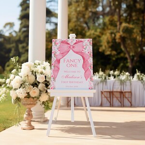 Op de afbeelding: Een roze en wit welkomsbord met een grote roze strik en bloemmotieven. Het bord staat er "Fancy One" en "Welkom op Madisons eerste verjaardag, 11 mei 20XX".