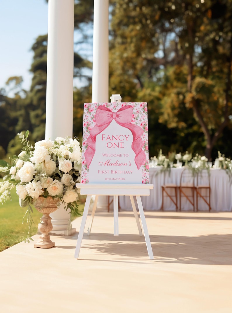 Op de afbeelding: Een roze en wit welkomsbord met een grote roze strik en bloemmotieven. Het bord staat er "Fancy One" en "Welkom op Madisons eerste verjaardag, 11 mei 20XX".