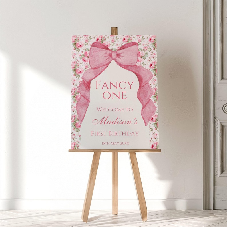 Op de afbeelding: Een roze en wit printbaar bord met een grote roze strik en de tekst "Fancy One"  "Welkom bij Madisons eerste verjaardag" en "15th May 20XX".