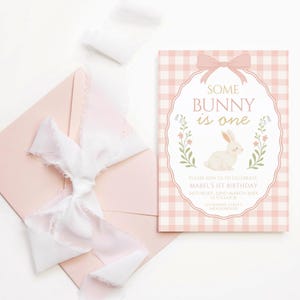 Op de afbeelding: Een roze en witte geruite uitnodigingskaart met de tekst "SOME BUNNY is one" en een konijnenillustratie. Een roze envelop en wit lint staan ook op de afbeelding. De uitnodiging is voor Mabel's 1e verjaardag.