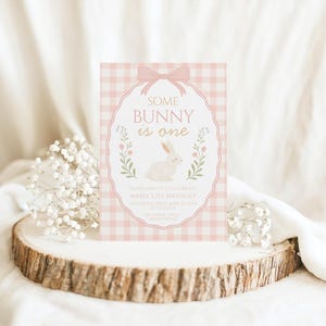 Op de afbeelding: Een roze en witte geruite uitnodigingskaart met de tekst "Some Bunny is One" en een witte konijnenillustratie. De uitnodiging is versierd met een roze strik en bloemaccenten. De kaart wordt gepresenteerd op een houten schijf met witte bloemen.