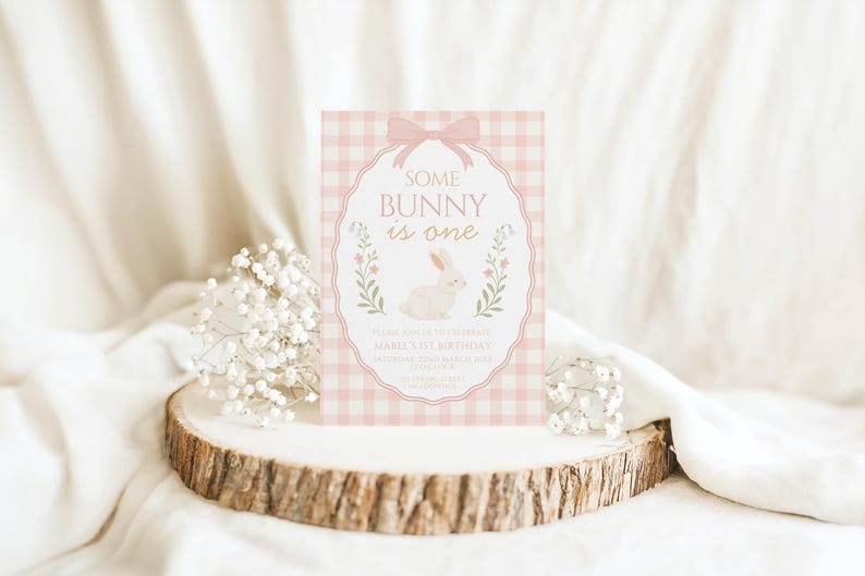 Op de afbeelding: Een roze en witte geruite uitnodigingskaart met de tekst "Some Bunny is One" en een witte konijnenillustratie. De uitnodiging is versierd met een roze strik en bloemaccenten. De kaart wordt gepresenteerd op een houten schijf met witte bloemen.