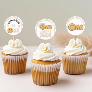 May include: Three cupcakes with white frosting and brown wrappers. Each cupcake has a round, brown and white paper topper with a smiley face and the text "One" or "Benjamin is One Happy Dude".