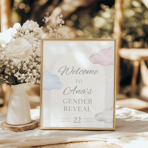 May include: A framed sign with gold trim, featuring the text "Welcome to Ana's Gender Reveal" with blue and pink watercolor clouds. The date "March 22" is displayed below. A white vase with white flowers and greenery sits to the left.