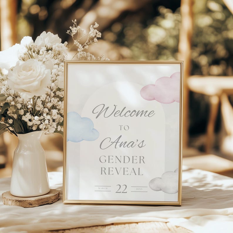 May include: A framed sign with gold trim, featuring the text "Welcome to Ana's Gender Reveal" with blue and pink watercolor clouds. The date "March 22" is displayed below. A white vase with white flowers and greenery sits to the left.