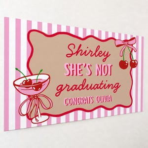 May include: A rectangular sign with a pink and white striped background. The sign features a brown shape with red trim and text that reads "Shirley SHE'S NOT graduating CONGRATS OLIVIA". A cocktail glass with cherries is on the left.