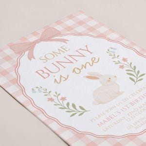 Op de afbeelding: Een roze en witte geruite uitnodiging met de tekst "Some Bunny is One". De uitnodiging bevat een aquarel illustratie van een konijn en bloemaccenten. De uitnodiging is voor Mabel's 1e verjaardag.