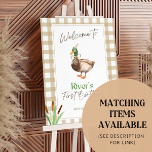 May include: Welcome to River's First Birthday party sign with a brown and green duck wearing a party hat and a bow. The sign has a brown and white gingham background with green reeds.