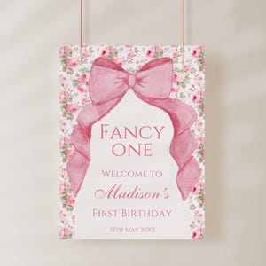 Op de afbeelding: Een roze en wit bloemenpatroonbord met een grote roze strik. Het bord zegt "Fancy One" en "Welkom op de eerste verjaardag van Madison 15 mei 20XX".