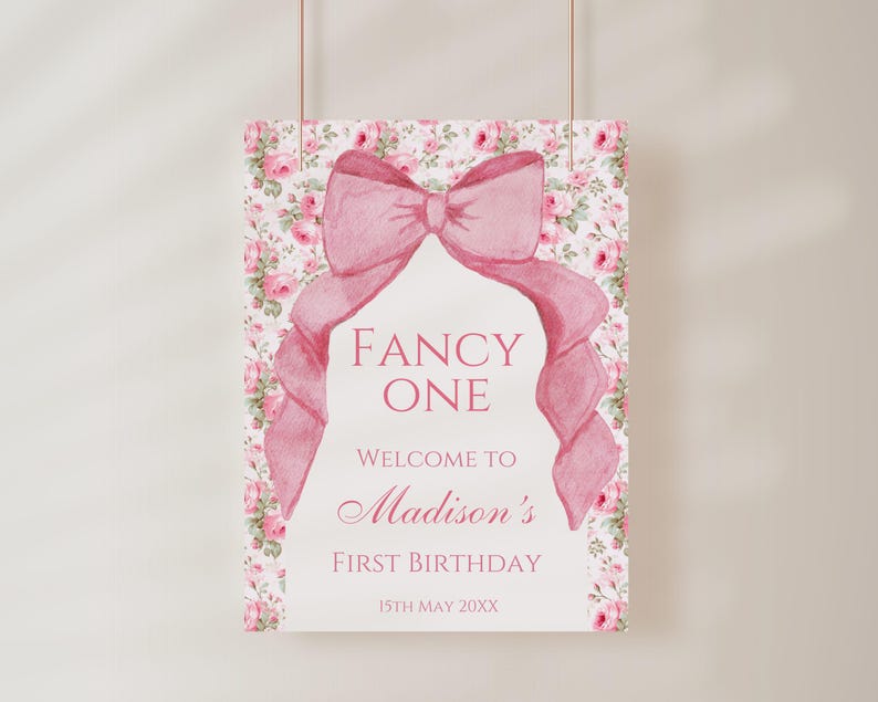 Op de afbeelding: Een roze en wit bloemenpatroonbord met een grote roze strik. Het bord zegt "Fancy One" en "Welkom op de eerste verjaardag van Madison 15 mei 20XX".
