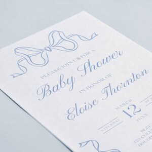 May include: A blue and white baby shower invitation with a blue ribbon bow at the top. The invitation reads "Please join us for a Baby Shower in honor of Eloise Thornton. Saturday, March 12, 20XX at 2 PM. Bleu Room, 123 Columbia Street, Boughville, CA 98765."