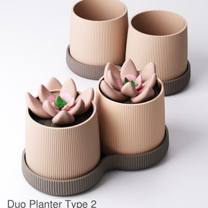 Op de afbeelding: Drie beige plantenbakken met een geribbelde textuur, elk op een donkergrijze basis. Twee zijn leeg, de derde bevat een kunstmatige vetplant met roze bloemblaadjes en een groen centrum. De tekst "Duo Planter Type 2" staat onderaan.