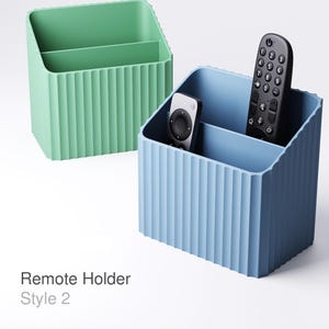 Può includere: Sono mostrati due porta telecomandi. Uno è verde chiaro, l'altro è azzurro. Ognuno ha due scomparti e un esterno a coste. Il supporto azzurro contiene due telecomandi neri. Il testo "Remote Holder Style 2" è in basso.