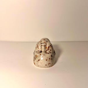 May include: A small ceramic sculpture of a human head with a glossy off-white finish and brown speckled spots. The face features closed eyes, a defined nose, and a small mouth, with the head tilted slightly upward in a neutral expression.