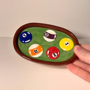 Può includere: Un vassoio ovale in ceramica con bordo marrone scuro e superficie interna verde. Il vassoio presenta cinque disegni circolari in rilievo di palle da biliardo in blu, bianco con viola, giallo, bianco con arancione e rosso, numerati 2, 12, 1, 13 e 3.