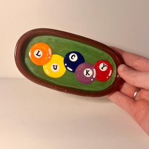 Op de afbeelding: Een ovaal keramieken dienblad met een donkerbruine rand en een groen binnenoppervlak. Het dienblad bevat vijf kleurrijke cirkelvormige ontwerpen die lijken op biljartballen, elk gemarkeerd met een zwarte letter om het woord LUCKY te spellen.