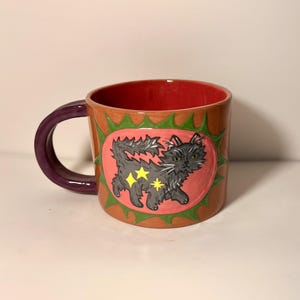 May include: A ceramic mug with a tan exterior and a red interior. The handle is purple. A central pink oval features a hand-painted black cat with yellow star accents, surrounded by a green spiked border. The mug has a smooth, glossy finish.