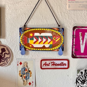 May include: A rectangular ceramic wall hanging featuring a stylized fish design in red, yellow, green, and white. The piece is framed by a yellow oval border on a blue background, suspended by a brown cord with two light blue teardrop charms.