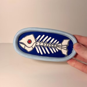 May include: An oval ceramic dish with a light blue rim and a deep blue center. The center features a white fish skeleton illustration with a single red dot for the eye. The dish has a glossy finish and a raised border around the central design.