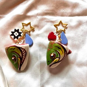 May include: A pair of earrings featuring hand-painted fish head charms in green, white, and red. Each earring includes a gold-tone star charm, a light blue teardrop, and one earring has a white square with a black star while the other has a red heart.