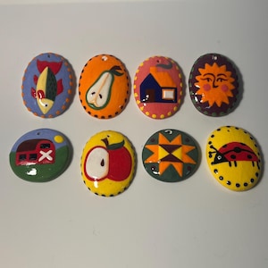 May include: Eight oval-shaped ceramic ornaments featuring hand-painted designs including a fish, a pear, a house, a sun, a barn, an apple, a geometric star pattern, and a ladybug. Each piece has a small hole at the top for hanging purposes.