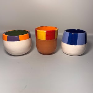 May include: Three ceramic tumblers with colorful, geometric designs. One has a purple and orange rim, another a yellow and red rim, and the third a blue rim. The lower portion of each tumbler is white.