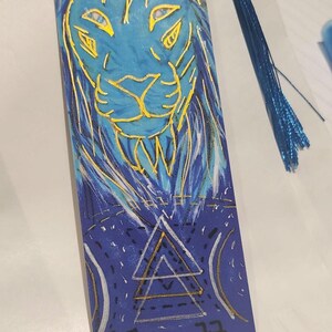 May include: A blue and gold bookmark with a lion's head design. The bookmark has a blue tassel attached at the top. The lion's face is outlined in gold, with a blue background. The bottom of the bookmark has a geometric design.