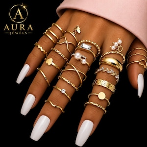 May include: A collection of gold-colored rings in various designs, including bands, twisted, and embellished with pearls and crystals. The rings are displayed on fingers with white nail polish, against a black background. The Aura Jewels logo is in the upper left corner.