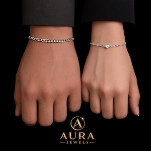 Puede incluir: Dos pulseras de cadena plateadas. Una es un diseño de cadena simple, la otra presenta un pequeño dije en forma de corazón. Las pulseras se muestran en muñecas sobre un fondo negro. El logotipo de Aura Jewels está en la parte inferior.