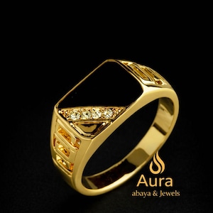 Op de afbeelding: Een goudkleurige ring met een rechthoekige zwarte steen en drie kleine heldere stenen. De ring heeft een decoratief ontwerp aan de zijkanten en staat tegen een zwarte achtergrond. De ring is van Aura abaya & Jewels.