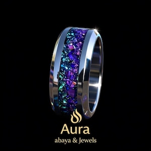 May include: A silver ring with a band of colorful, crushed gemstones in shades of purple, blue, and green. The ring has a polished, metallic finish and is set against a black background. The text "Aura abaya & Jewels" is at the bottom.