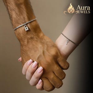 Può includere: Primo piano di due polsi con braccialetti d'argento. Uno presenta un piccolo ciondolo a forma di lucchetto, l'altro una chiave. Le mani sono intrecciate, simbolo di connessione. Il logo Aura Jewels è in alto a destra.