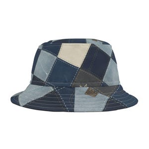 Op de afbeelding: Een blauwe bucket hat met een patchwork ontwerp. De hoed heeft vierkanten in verschillende tinten blauw, beige en grijs, aan elkaar gestikt met witte draad. De rand is afgewerkt met witte stiksels.