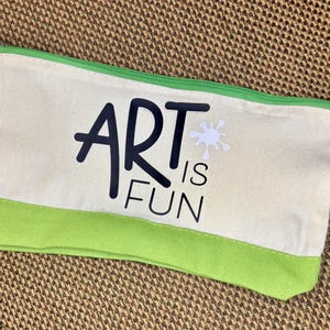 May include: A rectangular canvas pencil case featuring a cream-colored body with a bright green bottom panel and matching green zipper. The front displays the black text ART IS FUN with a small white paint splatter graphic above the word ART.