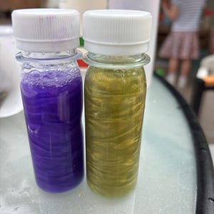 May include: Two clear plastic bottles with white screw-on caps, each filled with a swirling liquid. The left bottle contains a vibrant purple liquid with a metallic sheen, while the right bottle contains a shimmering gold liquid with visible swirls.