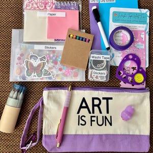 May include: A craft kit featuring a canvas pencil pouch with the text ART IS FUN, a dry erase board, a marker, washi tape, stickers, colored pencils, a stencil, and paper. The set includes various art supplies organized in clear plastic bags and boxes.