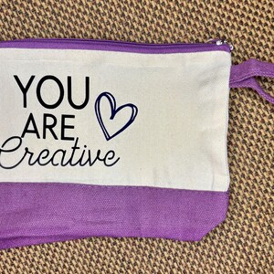 May include: A rectangular canvas zipper pouch featuring a cream-colored upper section and a purple lower section. The text "YOU ARE" is printed in black block letters above the word "Creative" in black script, next to a black heart outline.