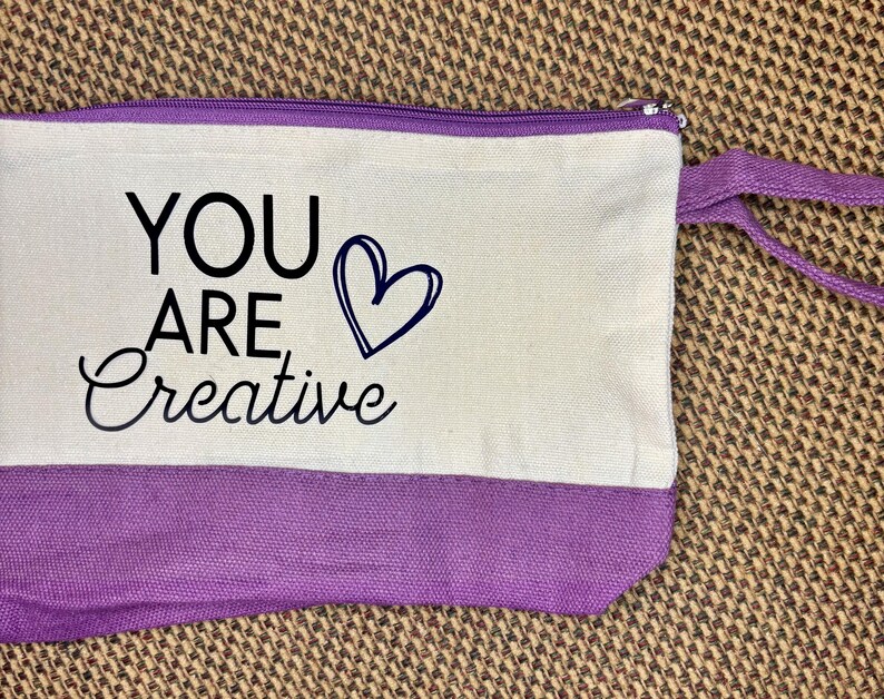 May include: A rectangular canvas zipper pouch featuring a cream-colored upper section and a purple lower section. The text "YOU ARE" is printed in black block letters above the word "Creative" in black script, next to a black heart outline.